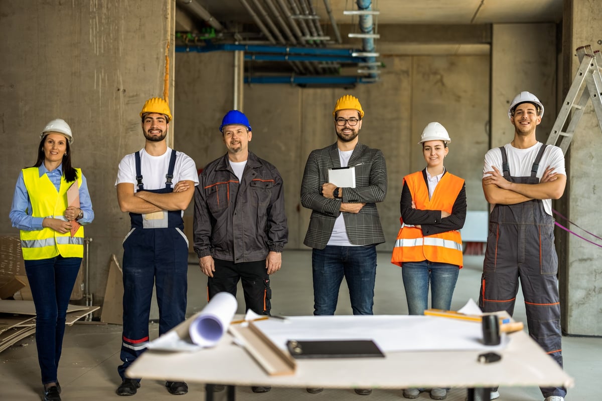 Proud construction crew with hard hats and safety vests smiling together. A work table with blueprints stands in front of them.