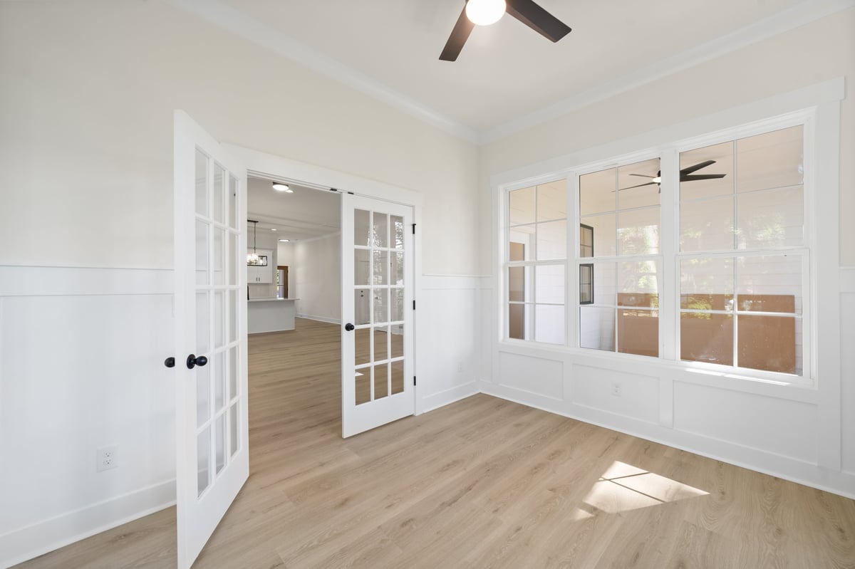 Empty bedroom with light wood flooring, white french doors, large windows with white frames, ceiling fan, and bright natural lighting