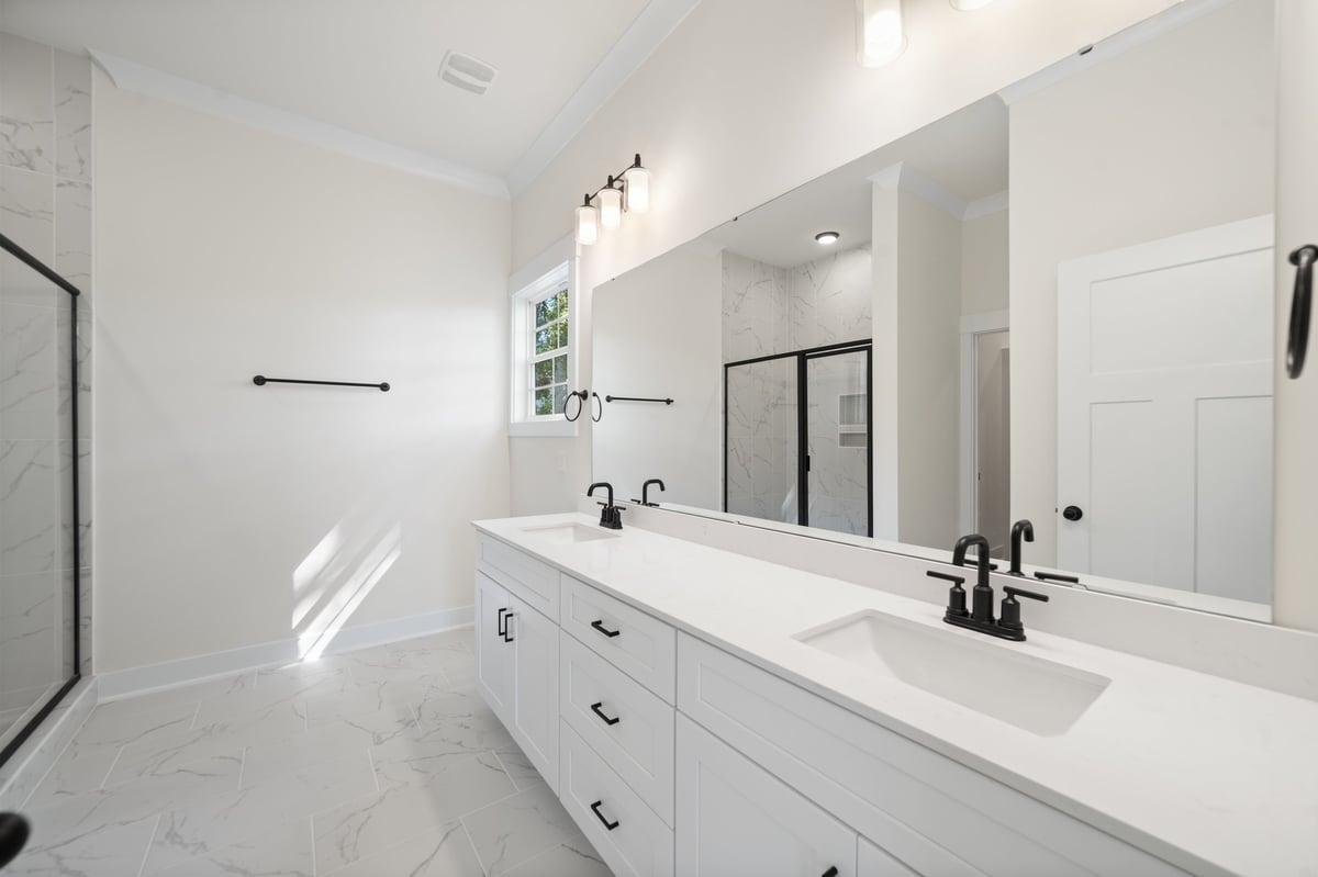 Modern white bathroom with double vanity sink, large mirror, and walk-in shower with glass door and marble flooring.