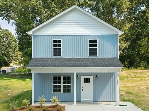 Two-story blue and white house with garage, two upper windows, front door with covered porch, surrounded by trees and green lawn