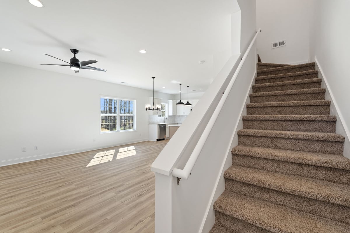 Modern interior with wooden staircase on right leading to upper floor, open-concept living room with light wood flooring, white walls, ceiling fan, and natural light from windows.