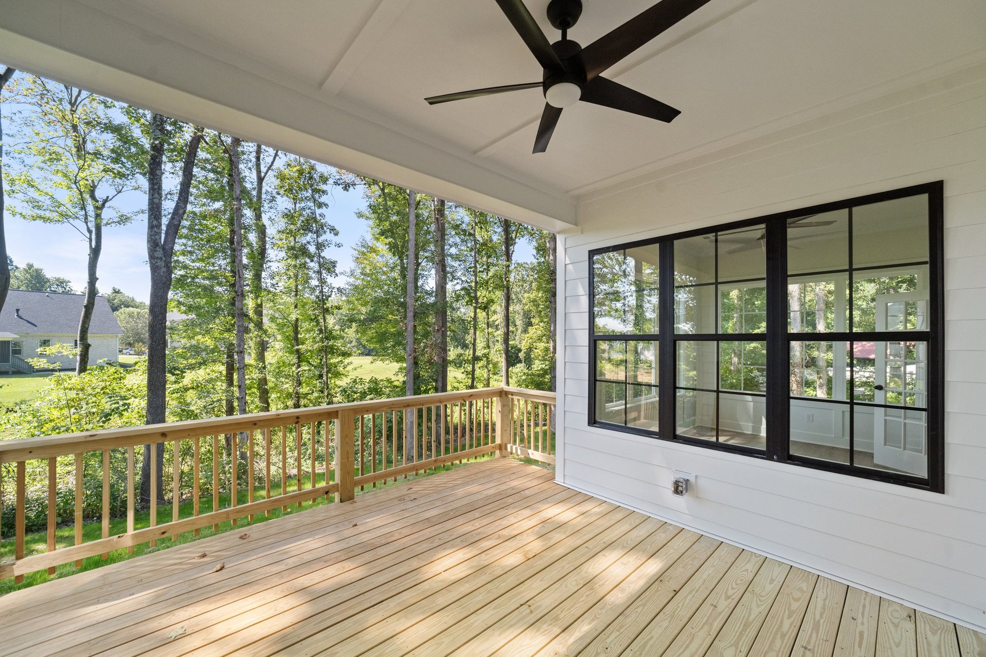 Covered wooden deck with railing, ceiling fan, and large black-framed windows overlooking green wooded landscape