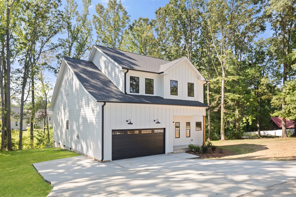 Modern white two-story house with black garage door and dormer, surrounded by tall trees on a sunny day