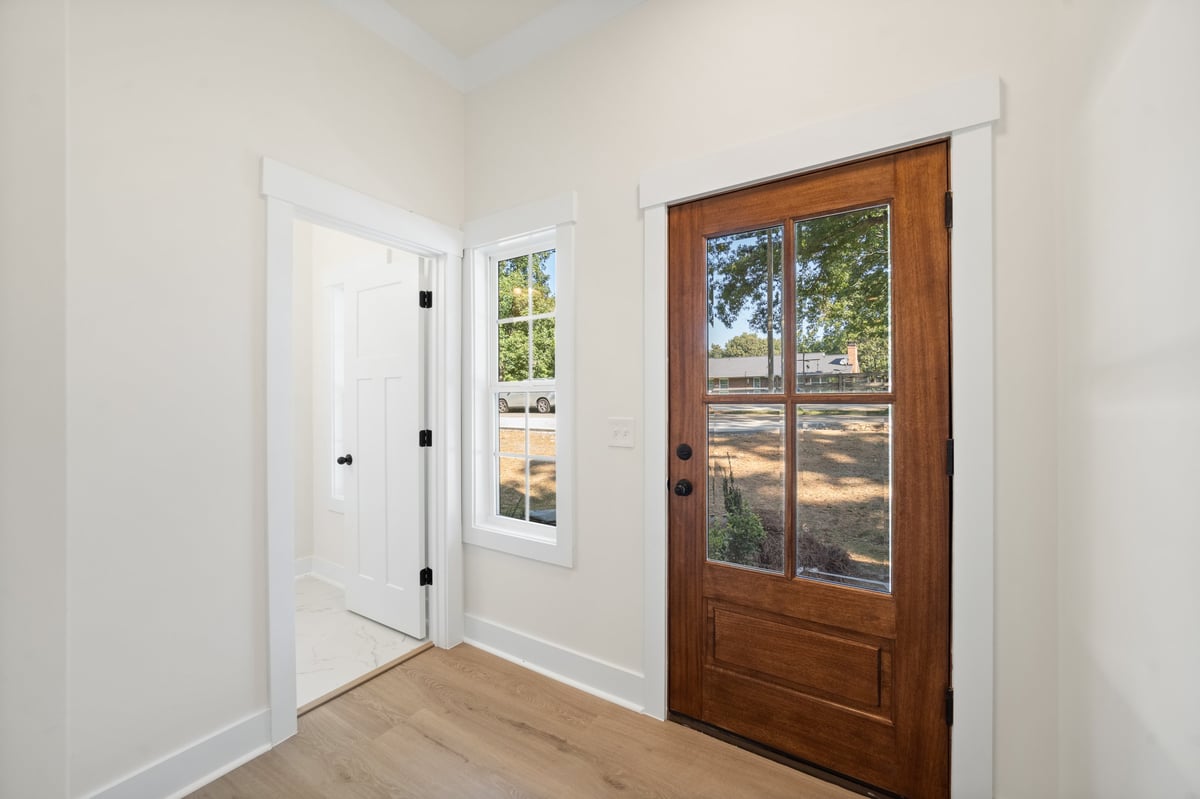 Modern entryway with white walls, wooden floors, a white interior door on the left, a transom window, and a wood-framed glass exterior door with tree views