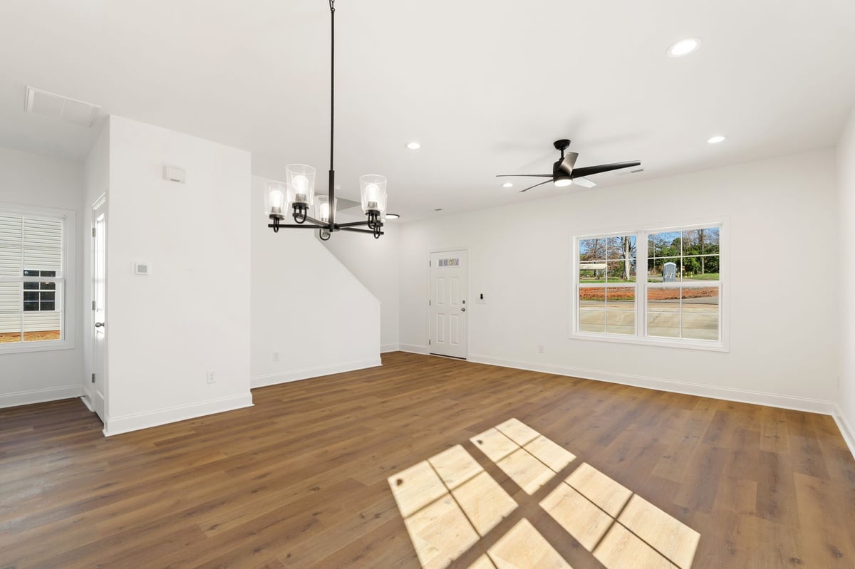 Empty modern living room with wood flooring, white walls, ceiling fan, pendant light, and windows with natural light casting shadows