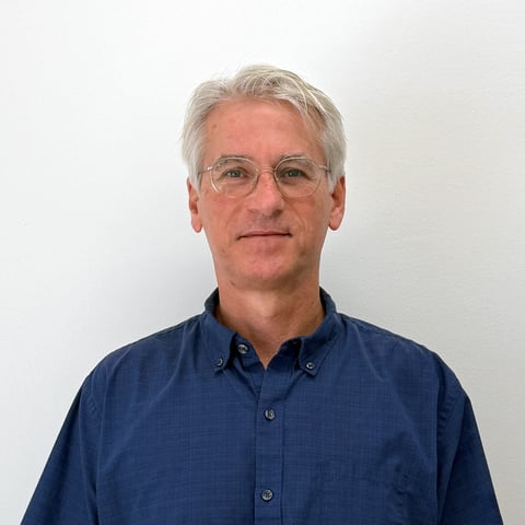 Middle-aged man with gray hair and glasses wearing a dark blue button-up shirt, portrait against white background