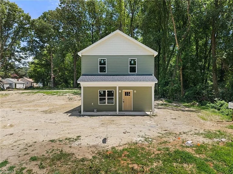 New beige two-story house under construction on bare lot surrounded by tall trees