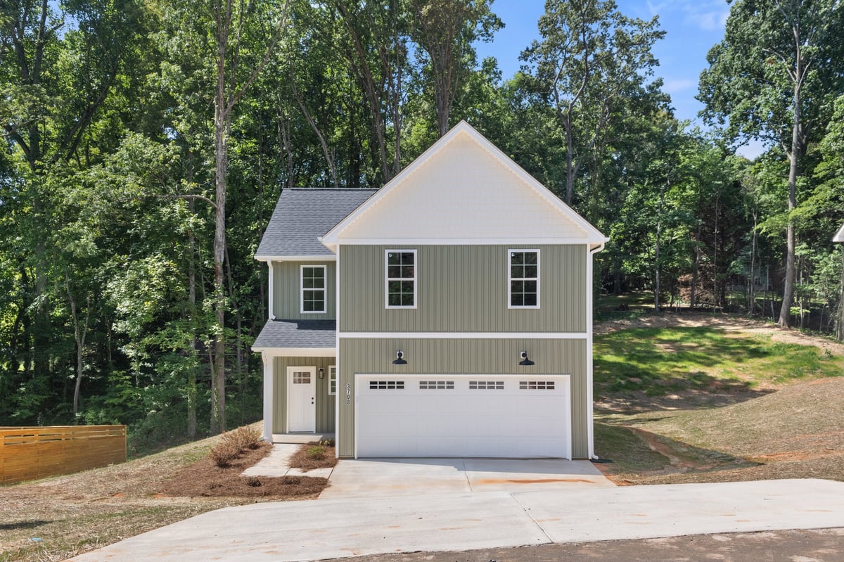 Modern two-story house with gray siding, white trim, and gabled roof, featuring a two-car garage and driveway surrounded by trees.