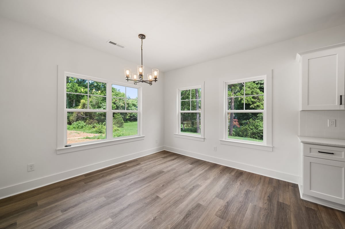Empty bright room with hardwood floors, white walls, three large windows with garden views, pendant light fixture, and white built-in cabinet.