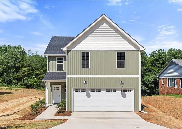 Two-story modern farmhouse with white garage door, tan siding, and pitched roof on a cleared lot surrounded by trees