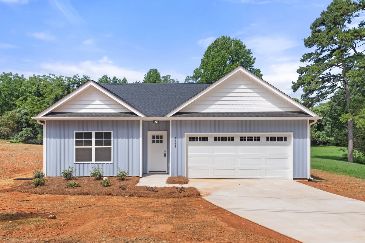 Modern white single-story house with dark roof, two-car garage, and driveway on red clay lot surrounded by green lawn and trees