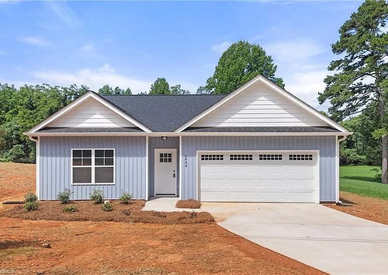 A single-story residential house with gray and white exterior, large garage, concrete driveway, and landscaped yard with trees.