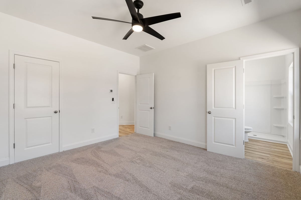 Empty bedroom with gray carpeting, white walls, black ceiling fan, and multiple white doors leading to closets and bathroom.