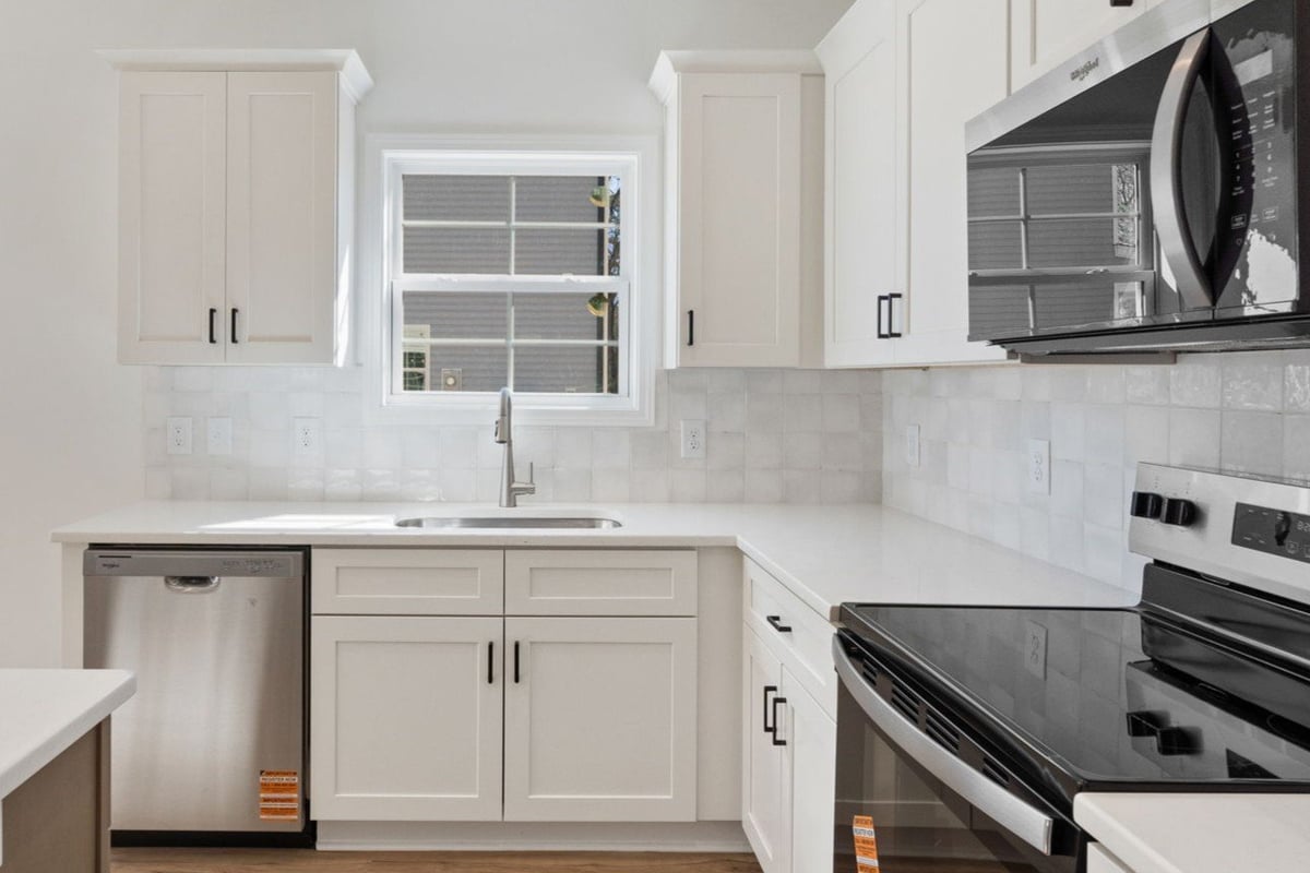 Modern white kitchen with stainless steel appliances, white cabinets, marble backsplash, and window above sink