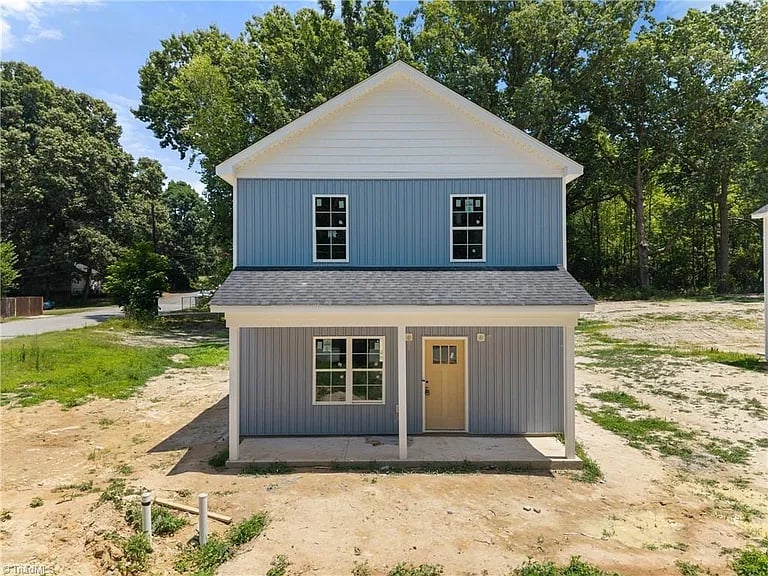 Two-story residential farmhouse with white and blue siding, wooden door, and windows on an unpaved lot surrounded by trees