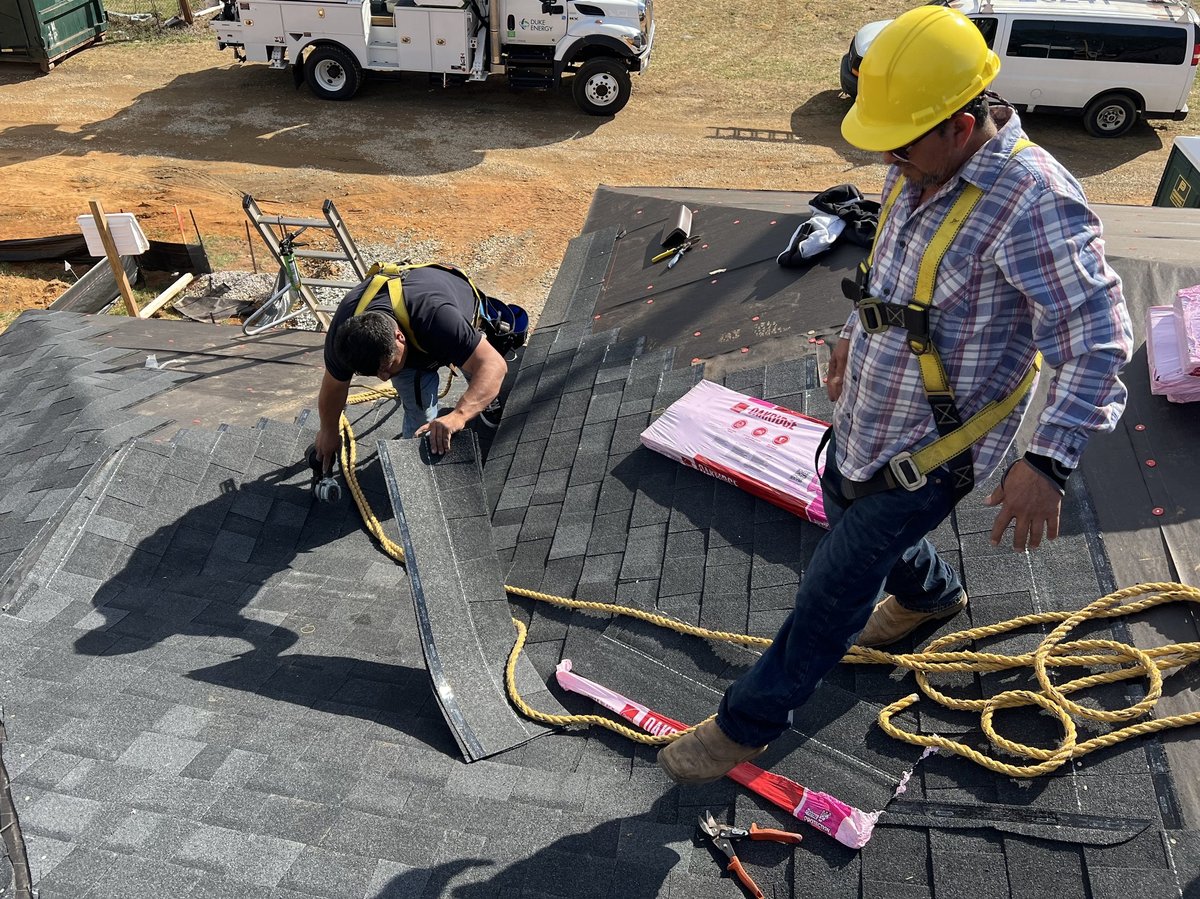 Two roofers installing shingles on a residential roof with service trucks and equipment visible in the background